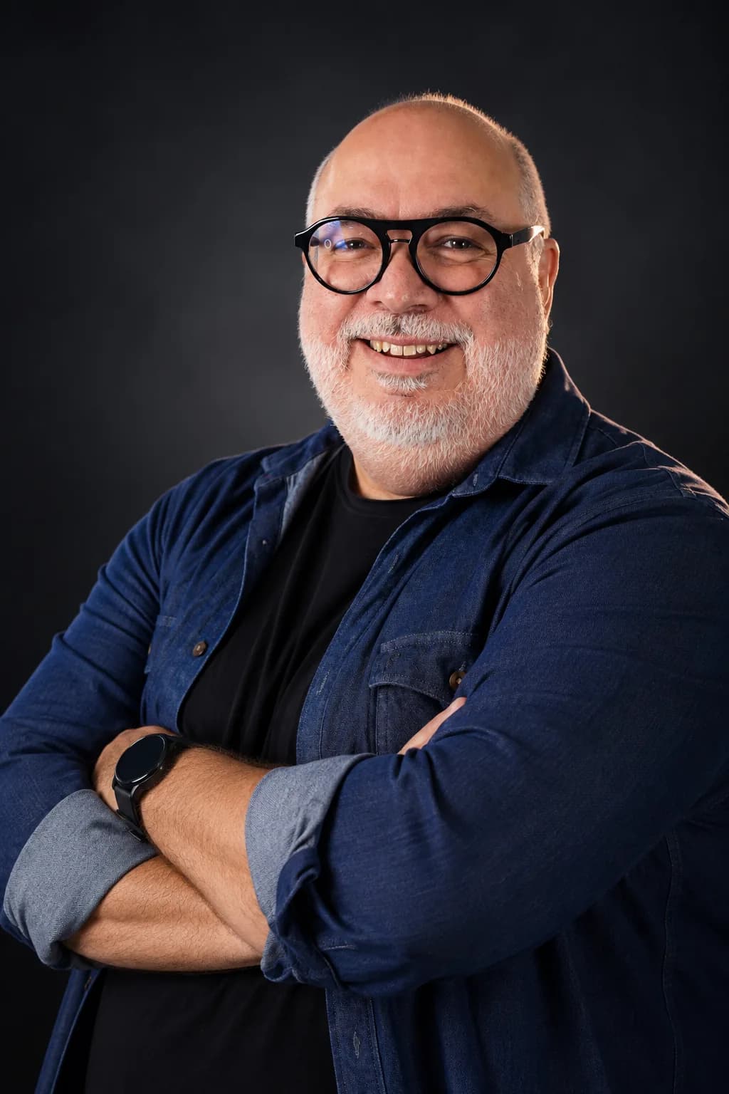 Retrato de Marcio Vitale, sorrindo, de braços cruzados, camisa jeans sobre camiseta preta, em fundo escuro.